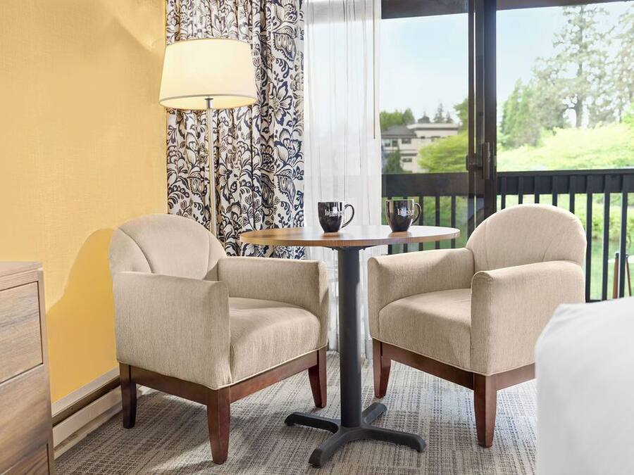 A hotel room with two armchairs, a small round table with a lamp, and a view of a balcony.