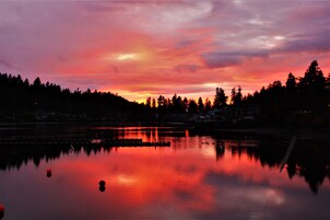 Atardecer sobre un lago tranquilo con reflejos, rodeado de árboles y una costa distante con edificios.