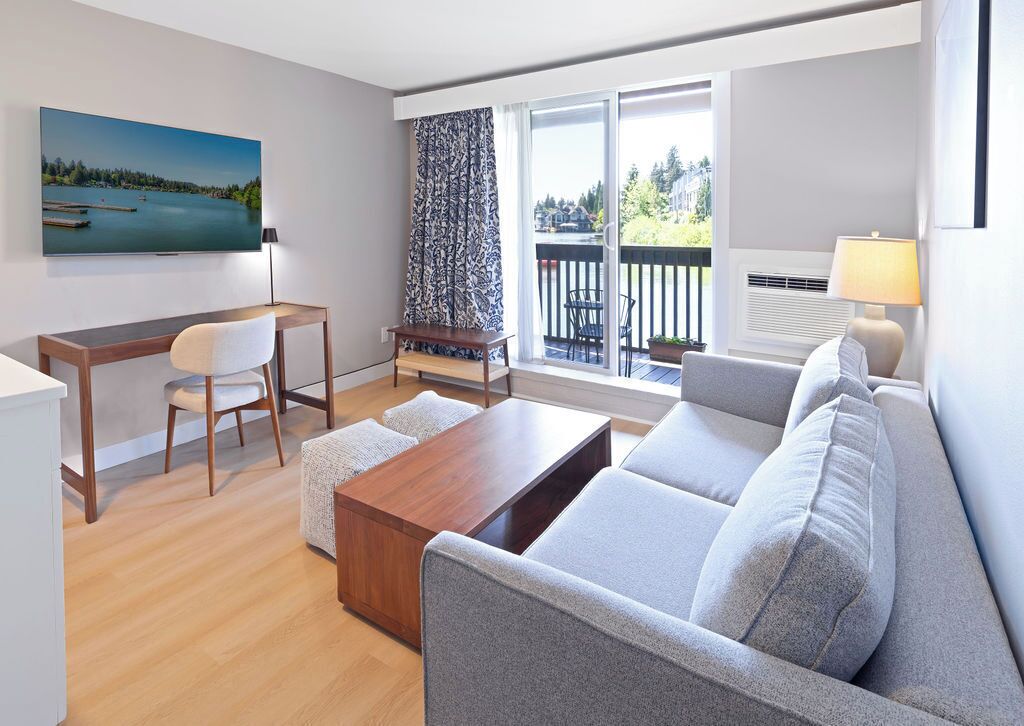A modern living room with a sofa, a coffee table, a TV, a desk with a chair, and a balcony with a view of the water.