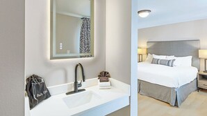 Hotellihuone, jossa on valkoinen pesuallas, peili, valkoisilla lakanoiden peitetty sänky, yöpöytä lampun kanssa ja pieni pyyhe sängyllä.