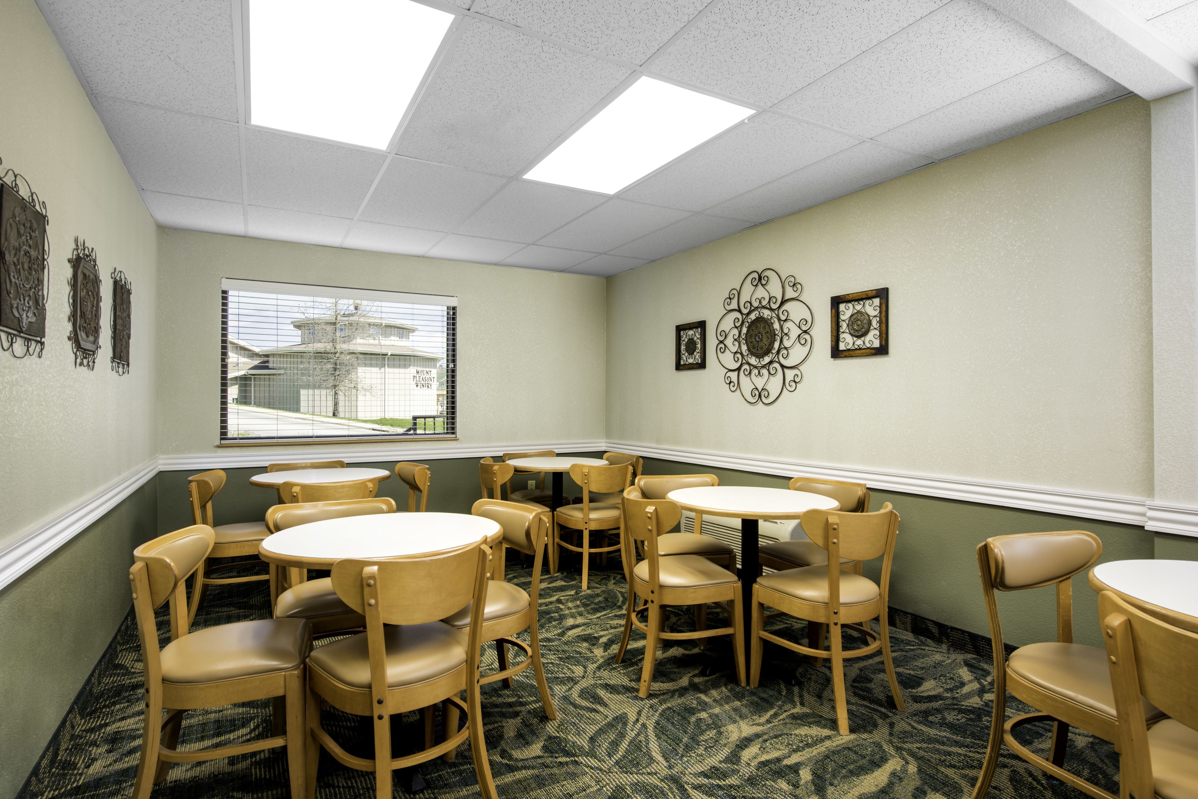 A hotel room with a dining area featuring round tables and wooden chairs, a window with a view of a building, and decorative wall art.