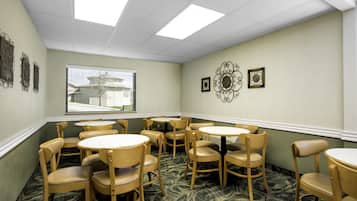 A hotel room with a dining area featuring round tables and wooden chairs, a window with a view of a building, and decorative wall art.