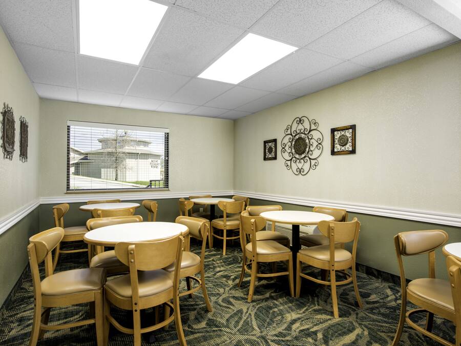 A hotel room with a dining area featuring round tables and wooden chairs, a window with a view of a building, and decorative wall art.