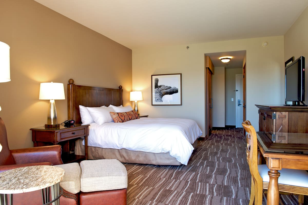 A hotel room with a large bed, two bedside tables with lamps, a wooden headboard, a flat-screen TV, a dining table with chairs, and a framed artwork on the wall.