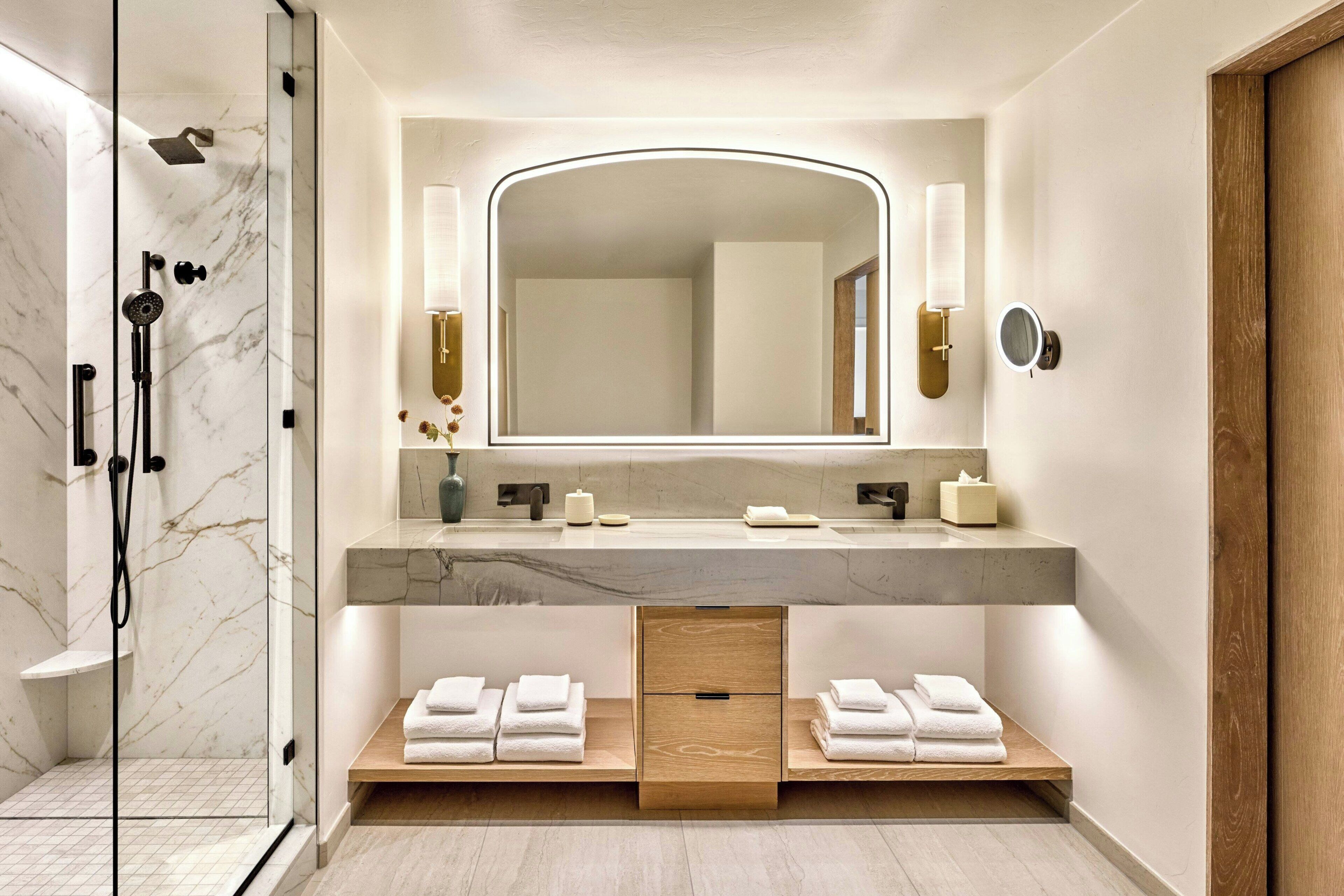 A modern bathroom with a glass-enclosed shower, a marble-tiled floor, a large mirror with wall sconces, and a wooden vanity with towels.