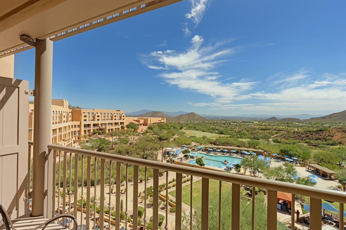 A balcony with a view of a resort pool area, including a large pool and lounge chairs, surrounded by greenery and mountains.