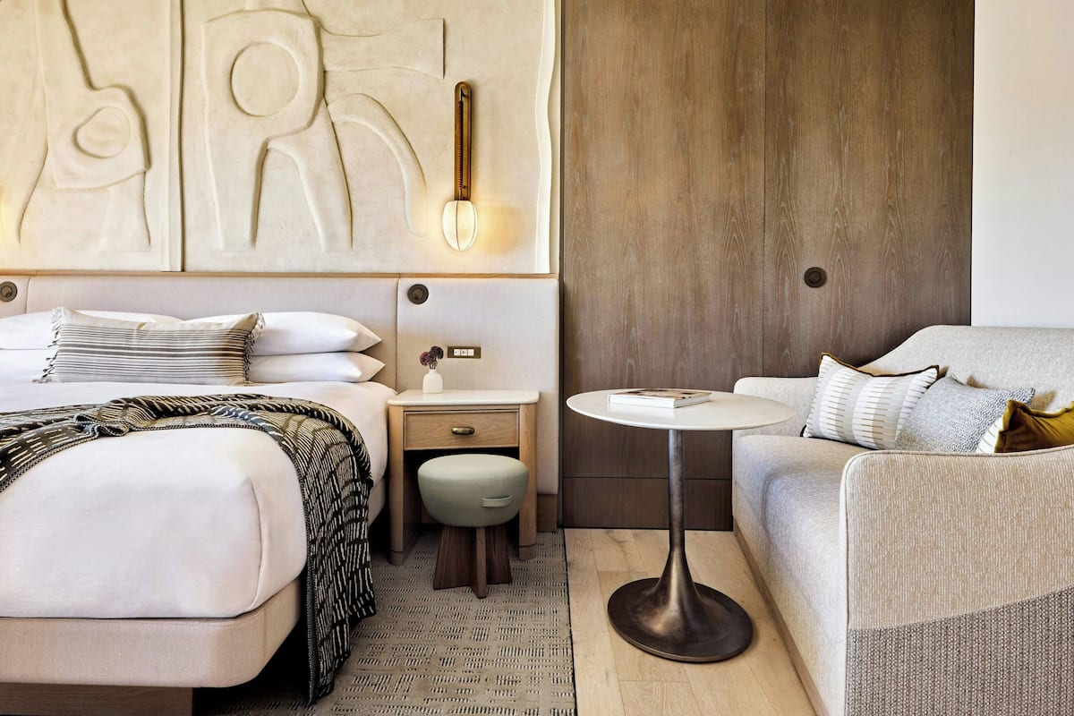 A modern hotel room with a large bed, a smaller sofa, a round table, and a decorative wall piece.