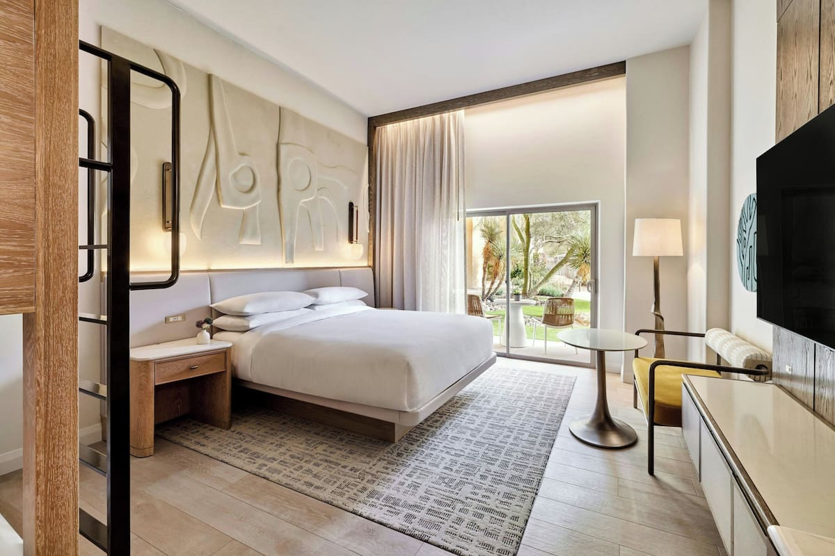 A modern hotel room with a large bed, a TV, a small table, and a balcony with a view.