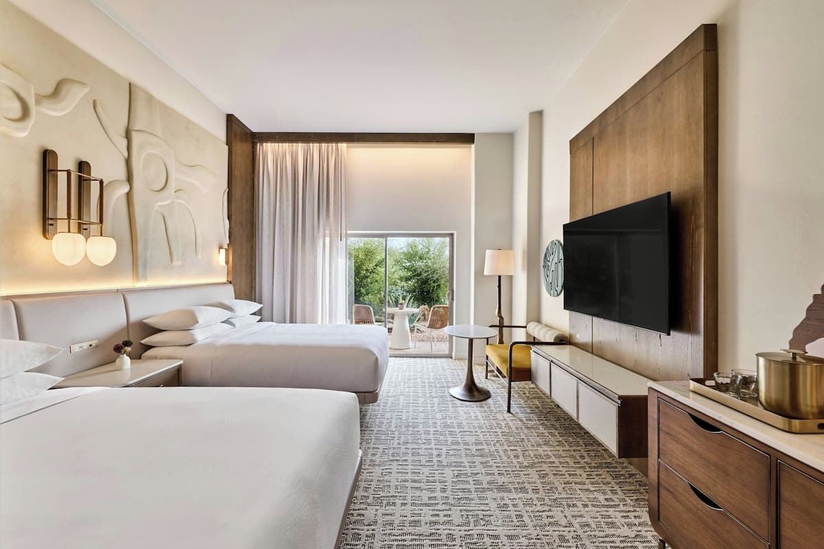 A hotel room with two beds, a TV, a dining area, and a view of the outdoors.