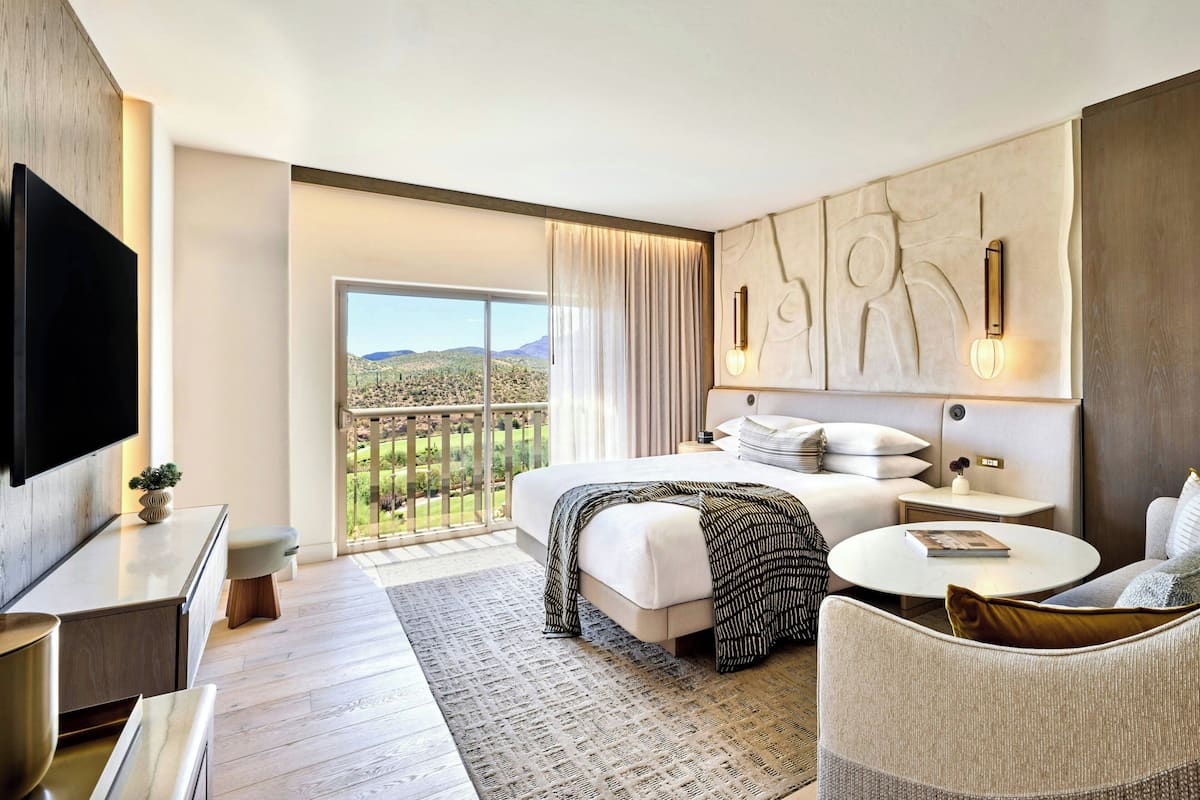 A modern hotel room with a large bed, a flat-screen TV, a balcony with a view of the mountains, and a round coffee table.