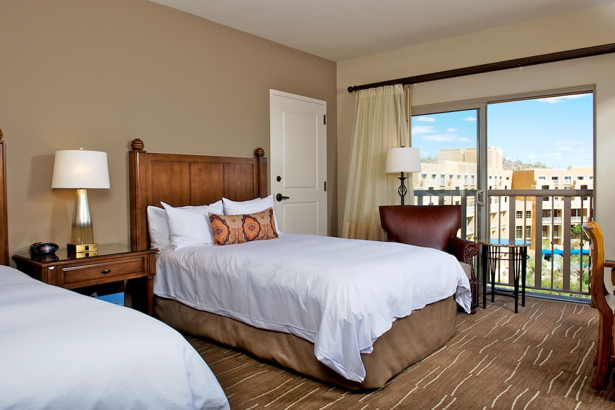 A hotel room with two beds, a wooden headboard, a nightstand with a lamp, a chair, and a balcony with a view of buildings.