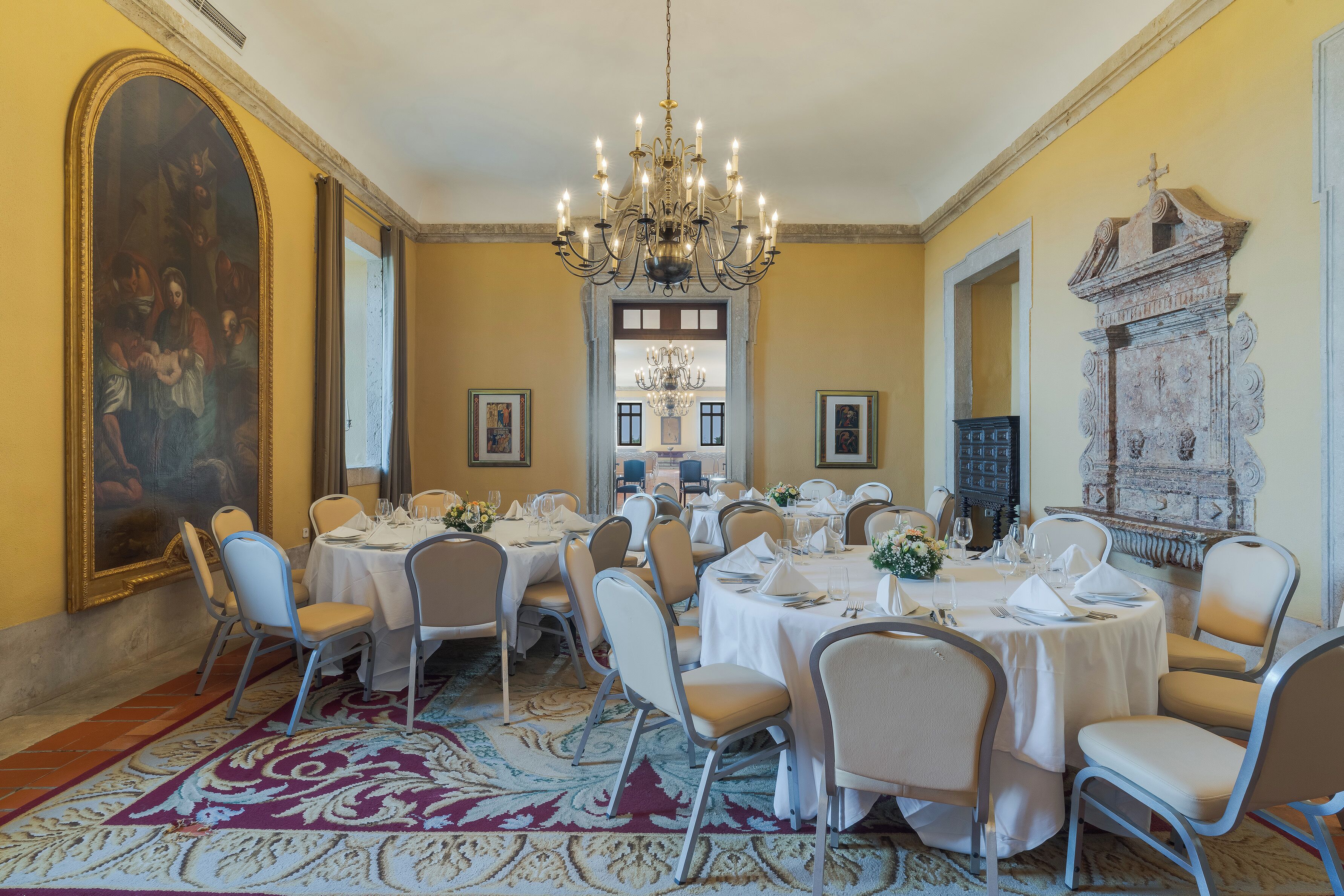 A dining room with round tables, chairs, a chandelier, and a large painting on the wall.