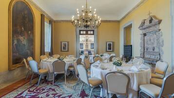 A dining room with round tables, chairs, a chandelier, and a large painting on the wall.