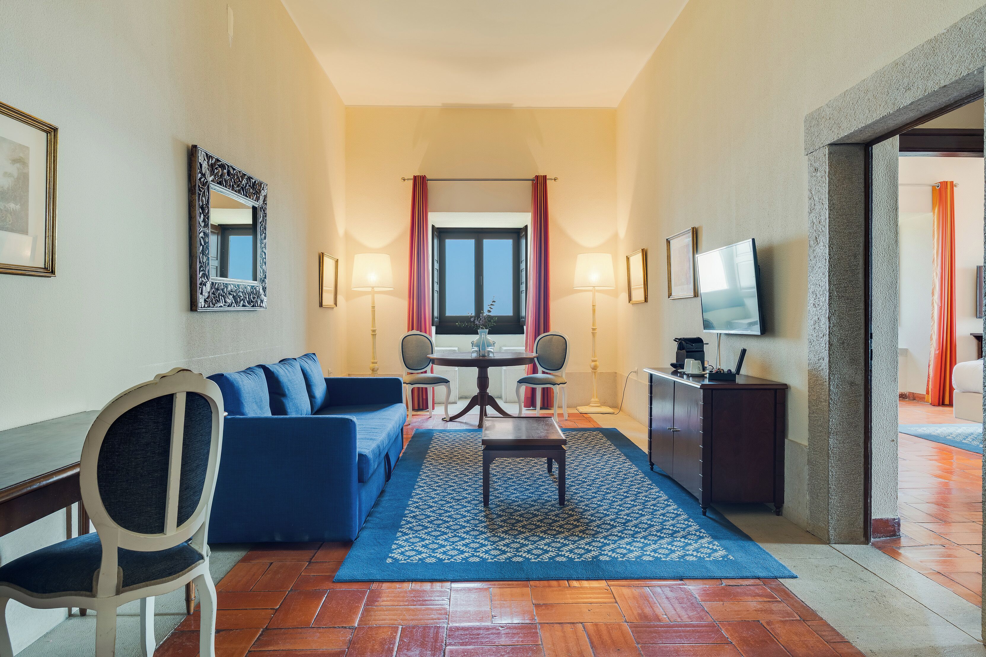 Une chambre d’hôtel avec un canapé bleu, une table à manger avec des chaises, un téléviseur et une grande fenêtre avec des rideaux.