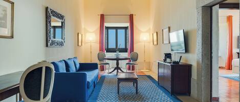 Une chambre d’hôtel avec un canapé bleu, une table à manger avec des chaises, un téléviseur et une grande fenêtre avec des rideaux.