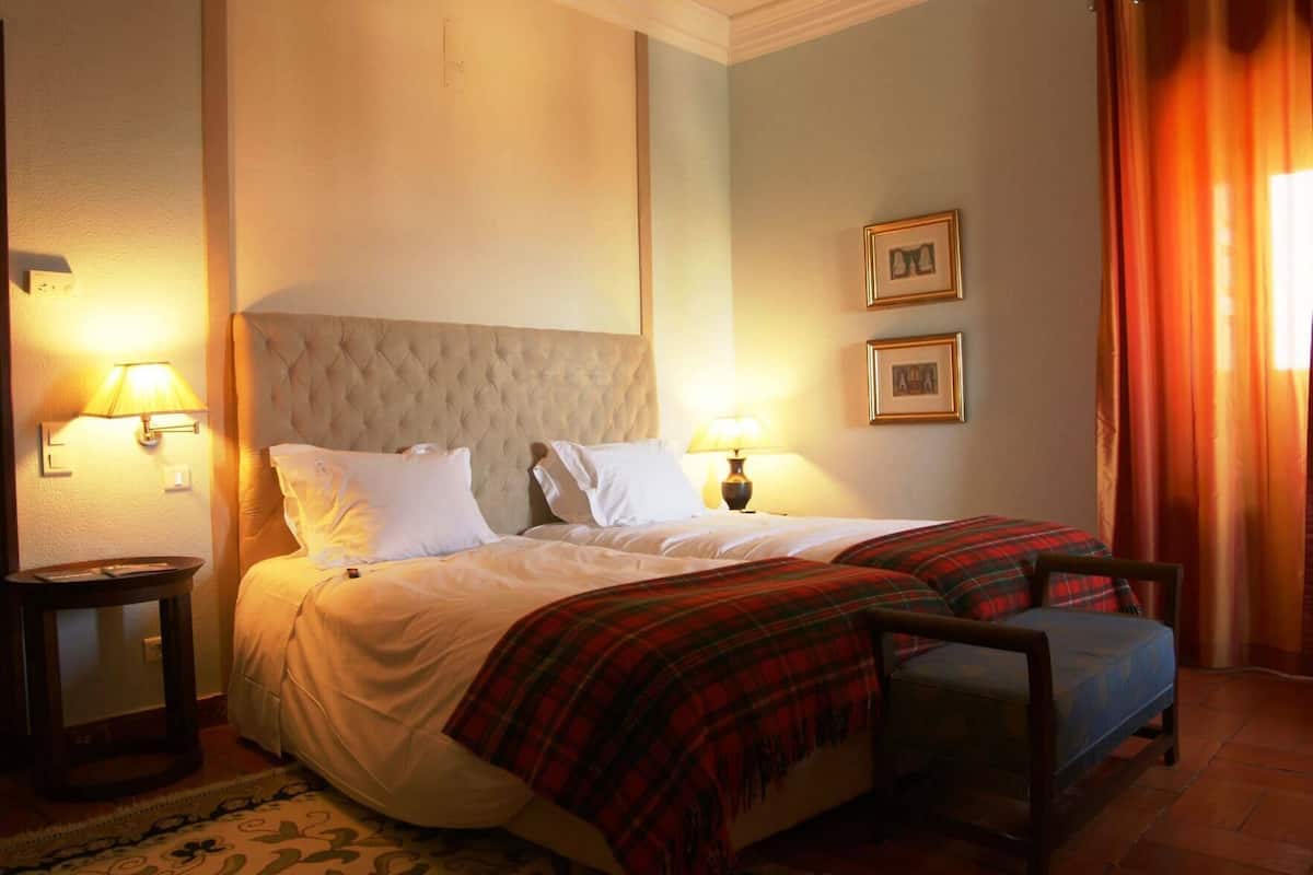 Habitación de hotel con una cama grande, dos mesitas de noche, una lámpara de noche, una colcha escocesa roja y dos cuadros en la pared.