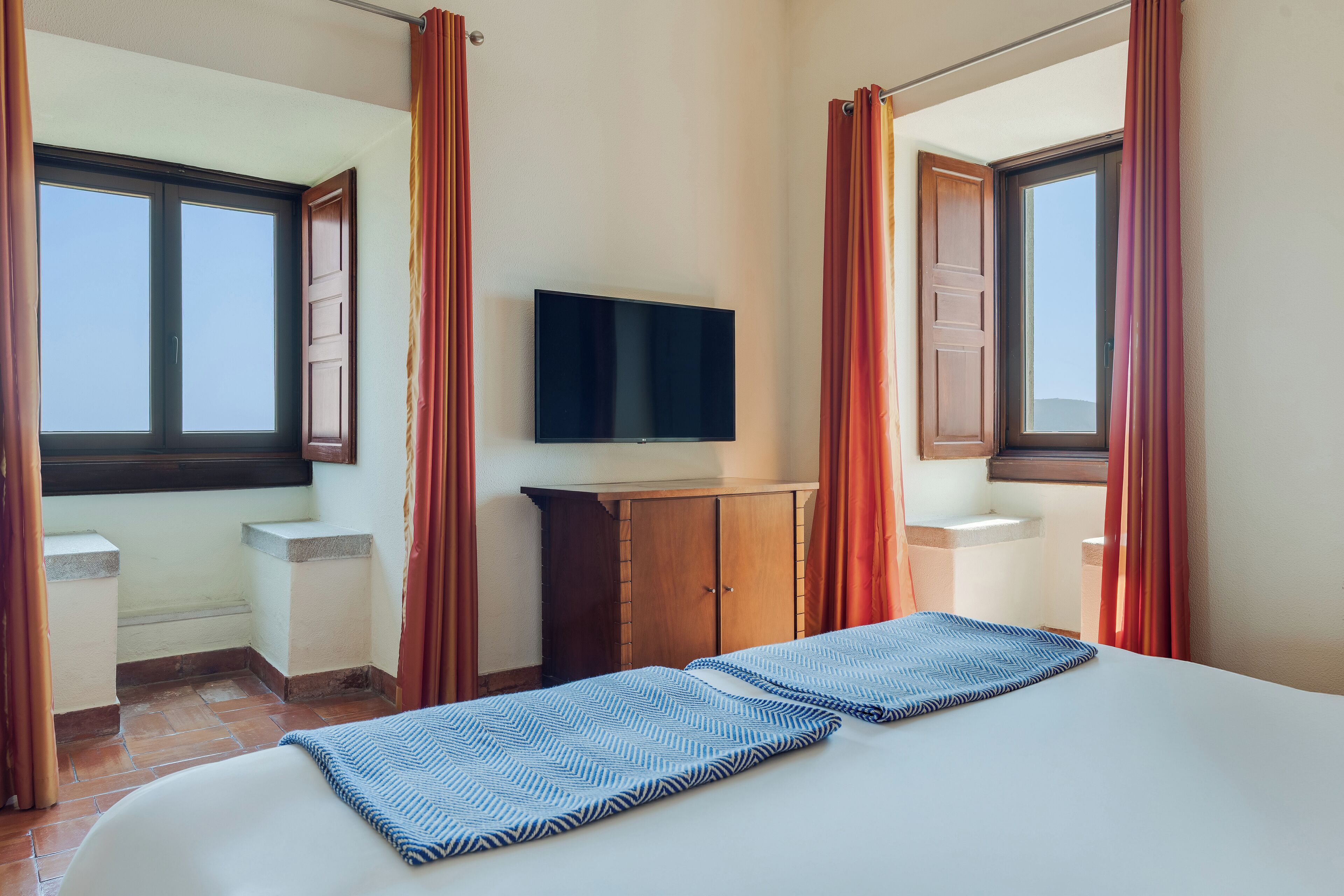 Quarto de hotel com cama, televisão sobre um móvel de madeira e janela com cortinas.