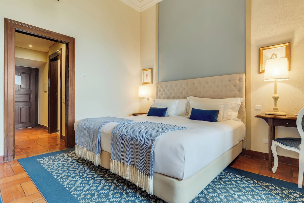 Un dormitorio ordenado con una cama grande, una mesita de noche, una silla y una puerta que da a otra habitación.