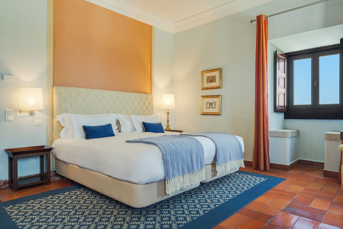 Un dormitorio ordenado con una cama grande, dos mesitas de noche con lámparas, una alfombra estampada y una ventana con cortinas.