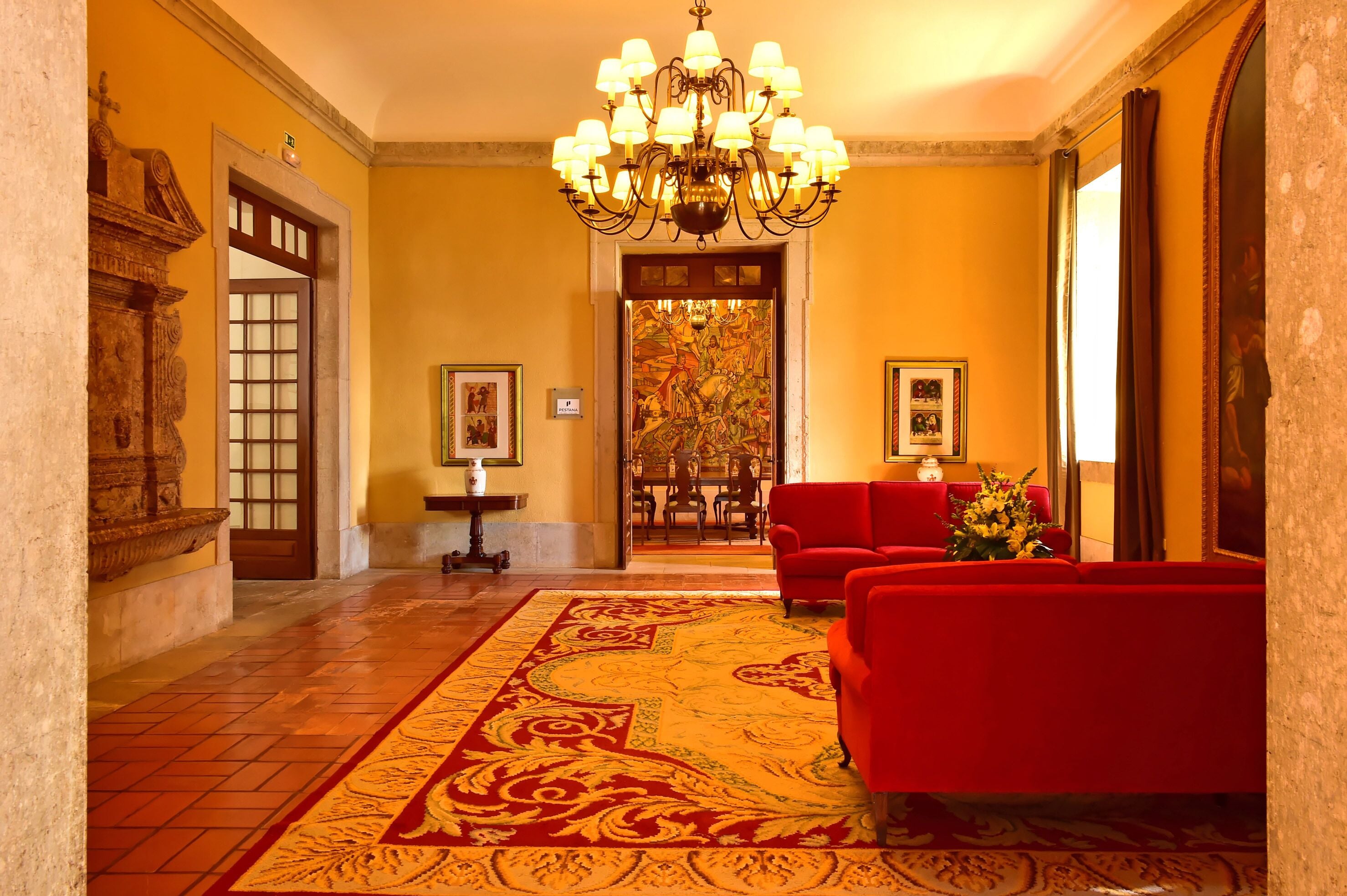 A lobby with a chandelier, red sofas, a patterned carpet, and a door leading to a dining area.
