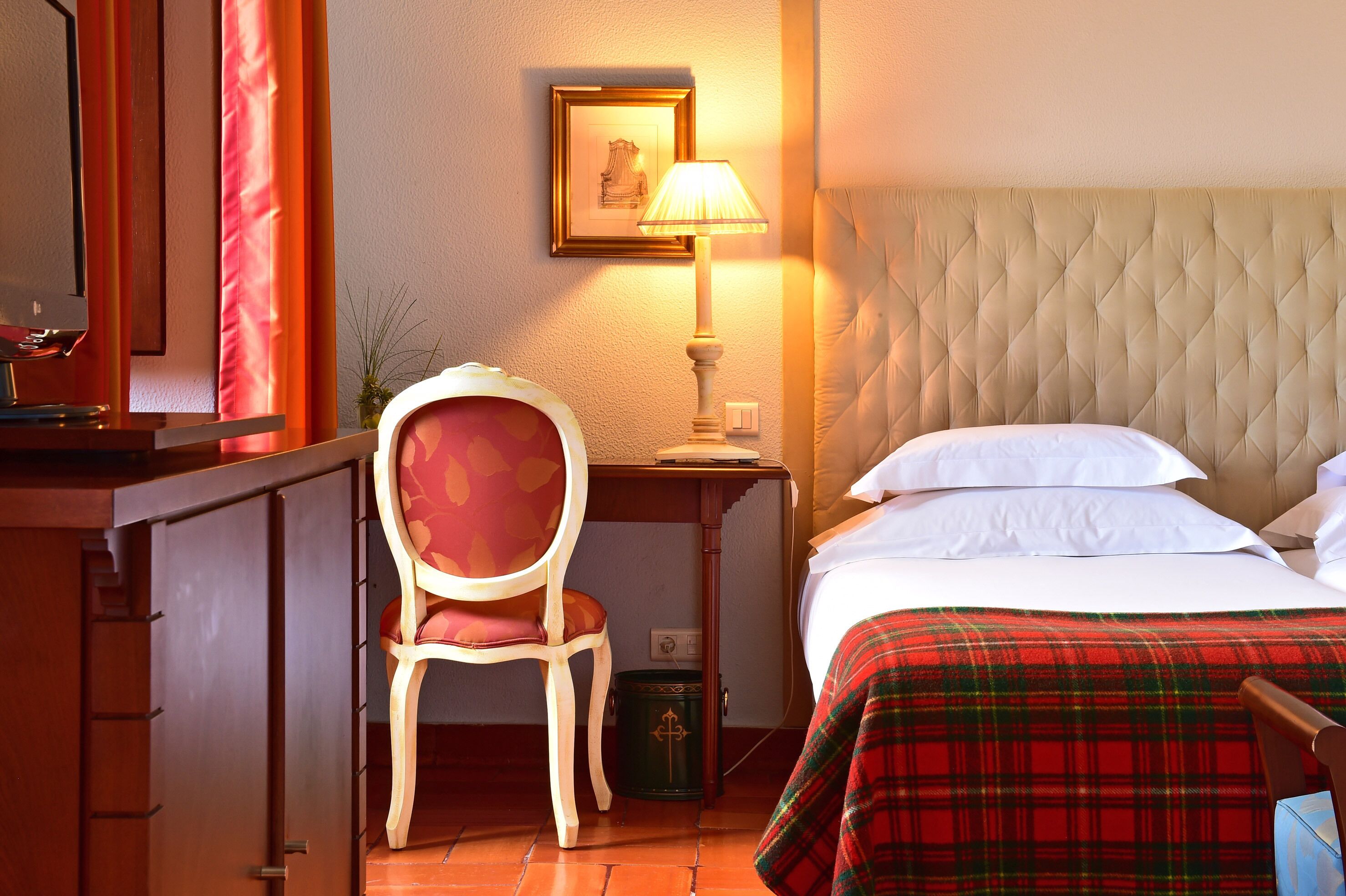 A hotel room with a bed, a chair, a desk, a lamp, and a framed picture on the wall.