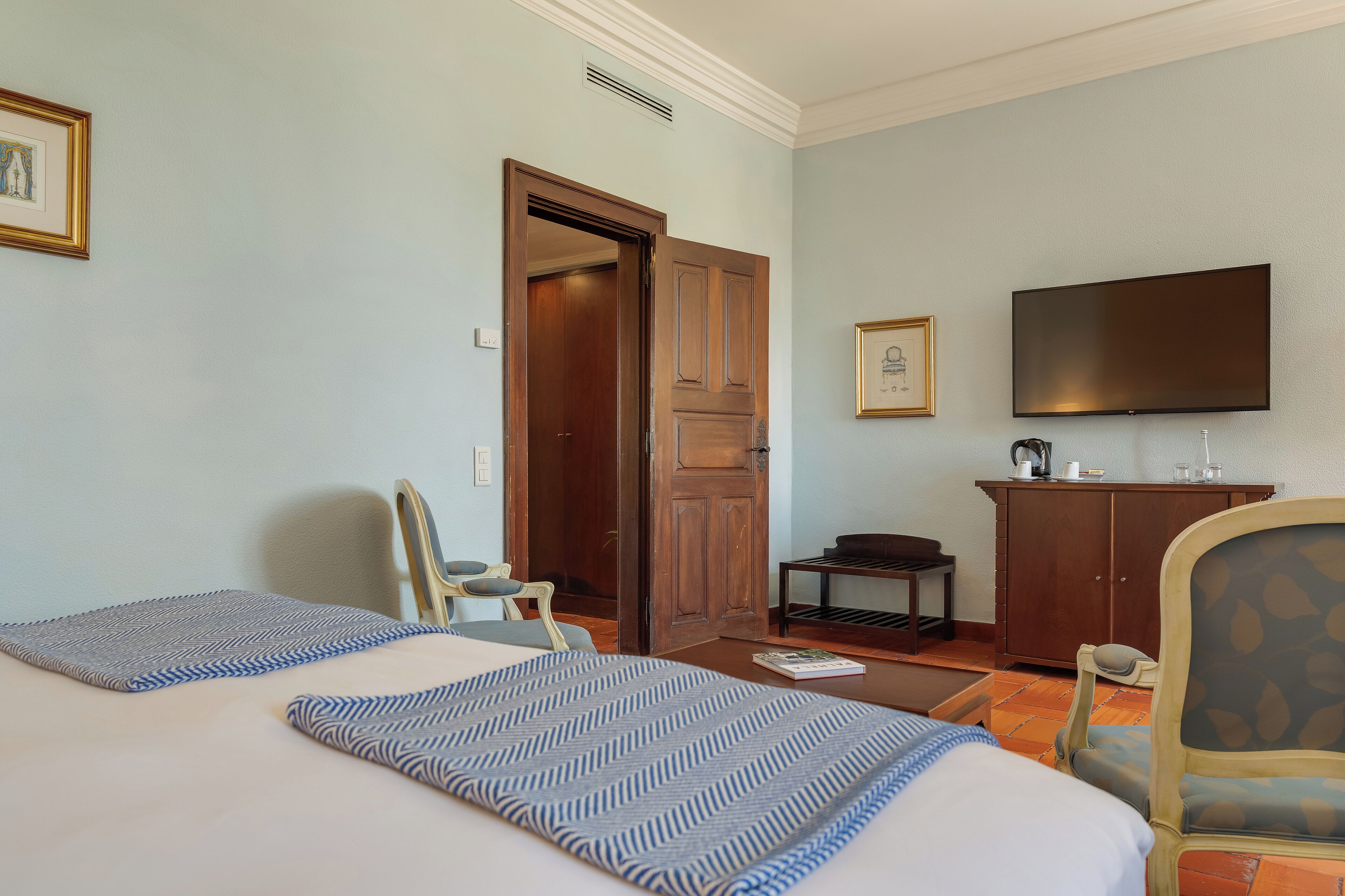 A hotel room with two beds, a television, a wooden wardrobe, and a small table.