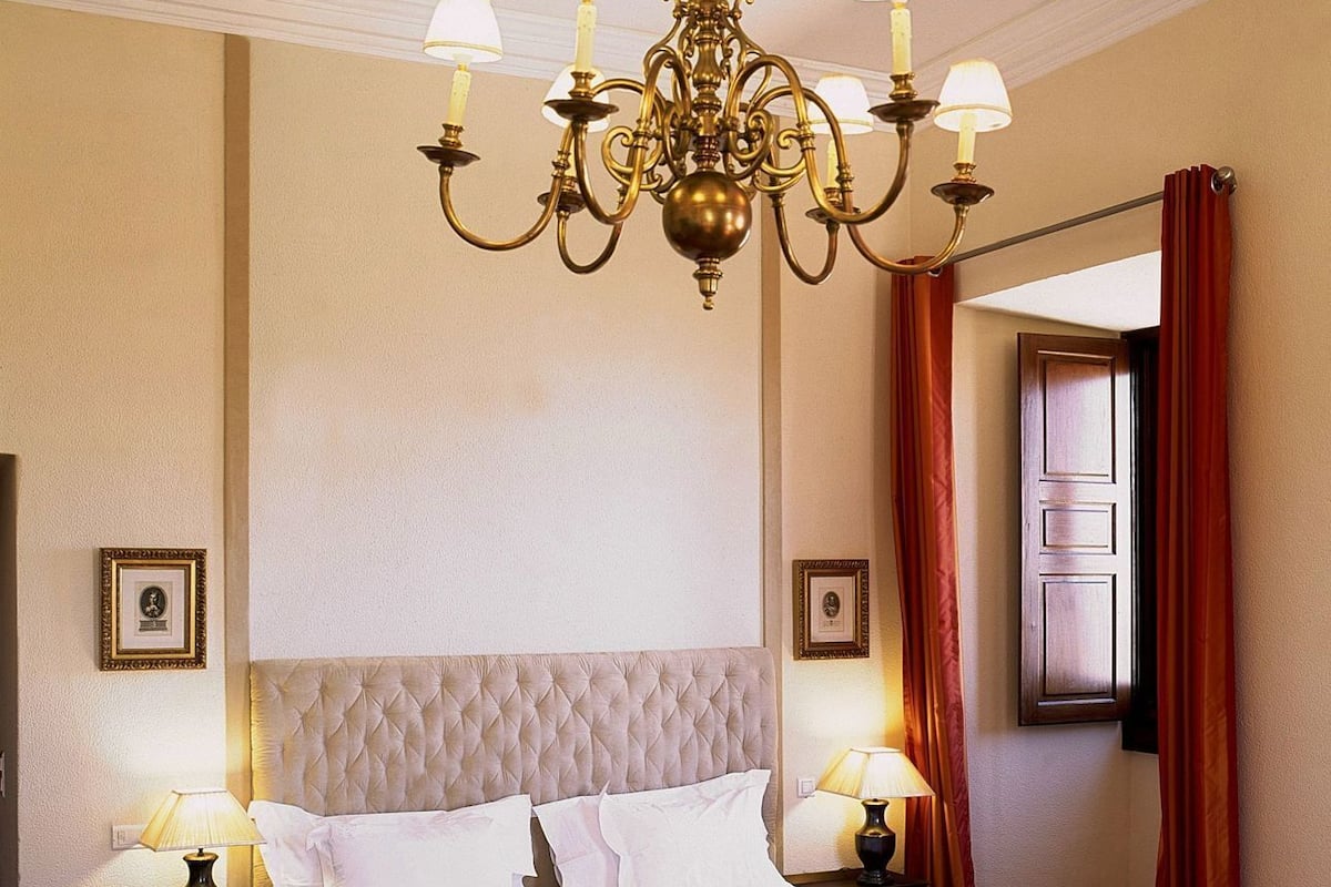 Una habitación de hotel con una cama grande, un candelabro, dos mesitas de noche con lámparas y una ventana con cortinas.