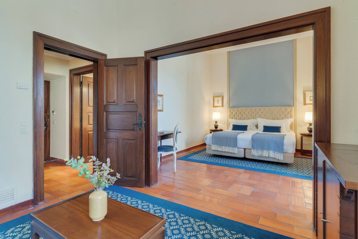 Una habitación de hotel con puerta de madera, una cama con cabecera azul, una mesita de noche, una lámpara, una silla y un florero con flores sobre una mesa.