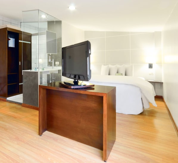 A modern hotel room with a wooden desk, a flat-screen TV, a bed with white linens, and a glass shower enclosure.