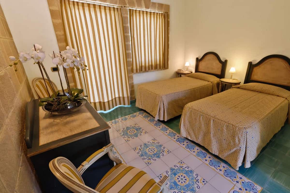 A hotel room with two beds, a desk with a chair, a small table with a lamp, and a window with striped curtains.