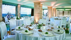 Un espace repas avec des tables rondes dressées pour le repas, des chaises blanches et une vue sur la mer grâce à de grandes fenêtres.