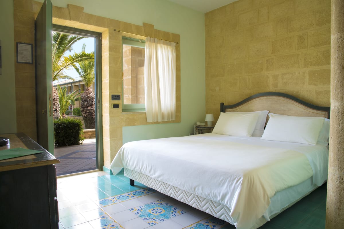 A bedroom with a bed, a nightstand, a window with curtains, and a view of palm trees outside.