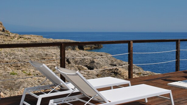 Une terrasse en bois, agrémentée de chaises longues blanches et d’un parasol blanc, offre une vue sur une côte rocheuse et la mer.