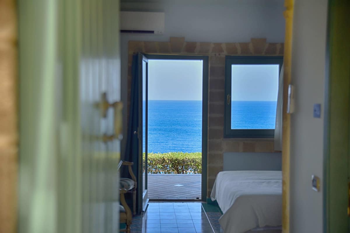 A room with a bed, a window with a view of the sea, and a door leading to another room.