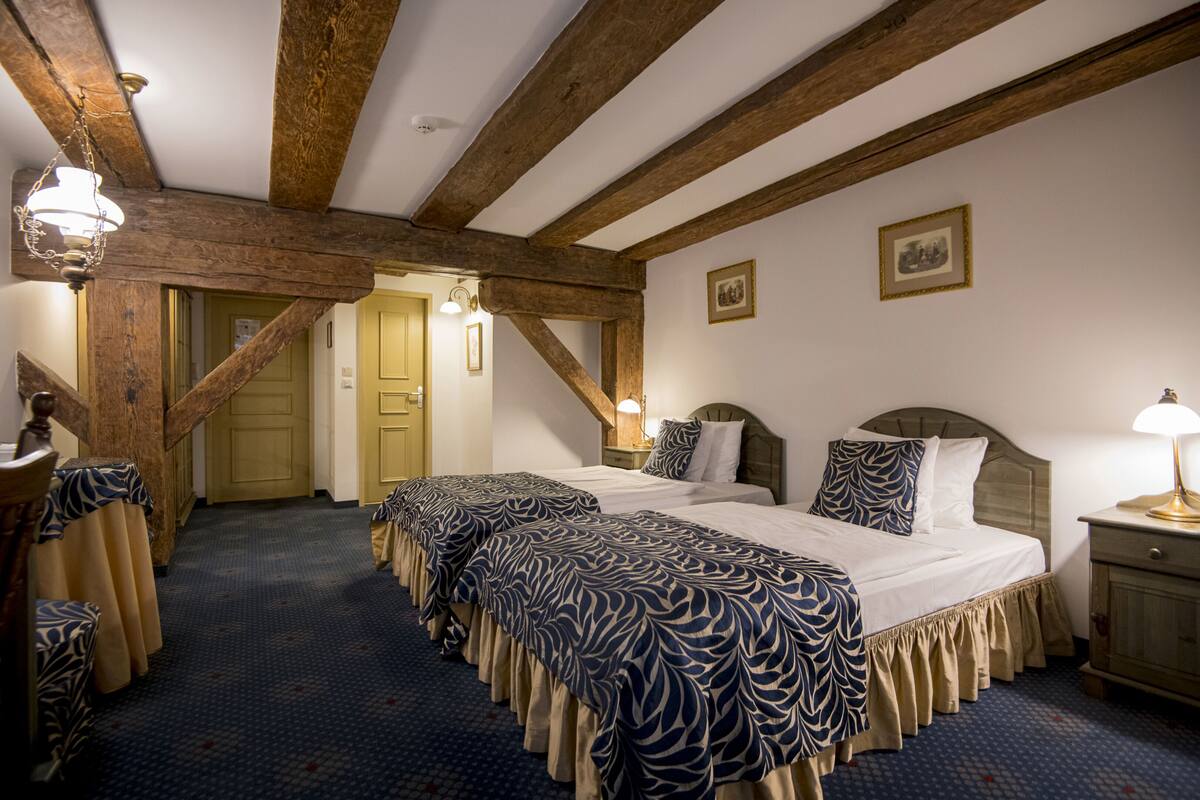 A hotel room with two beds, wooden beams, a patterned carpet, and a small table with a lamp.