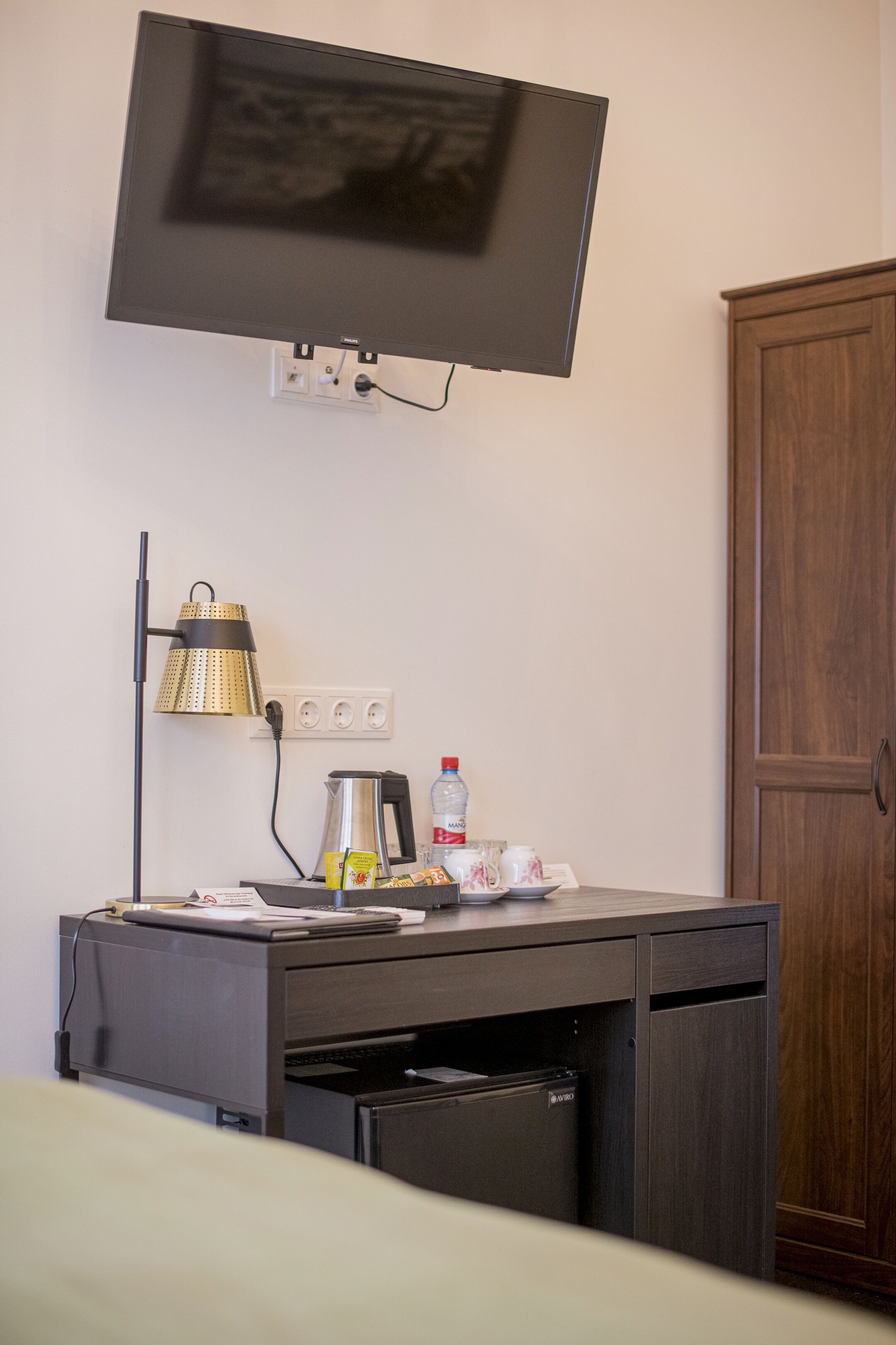 Hotellihuone, jossa on seinälle asennettu taulutelevisio, työpöytä lampun kanssa, vedenkeitin ja pullo vettä.