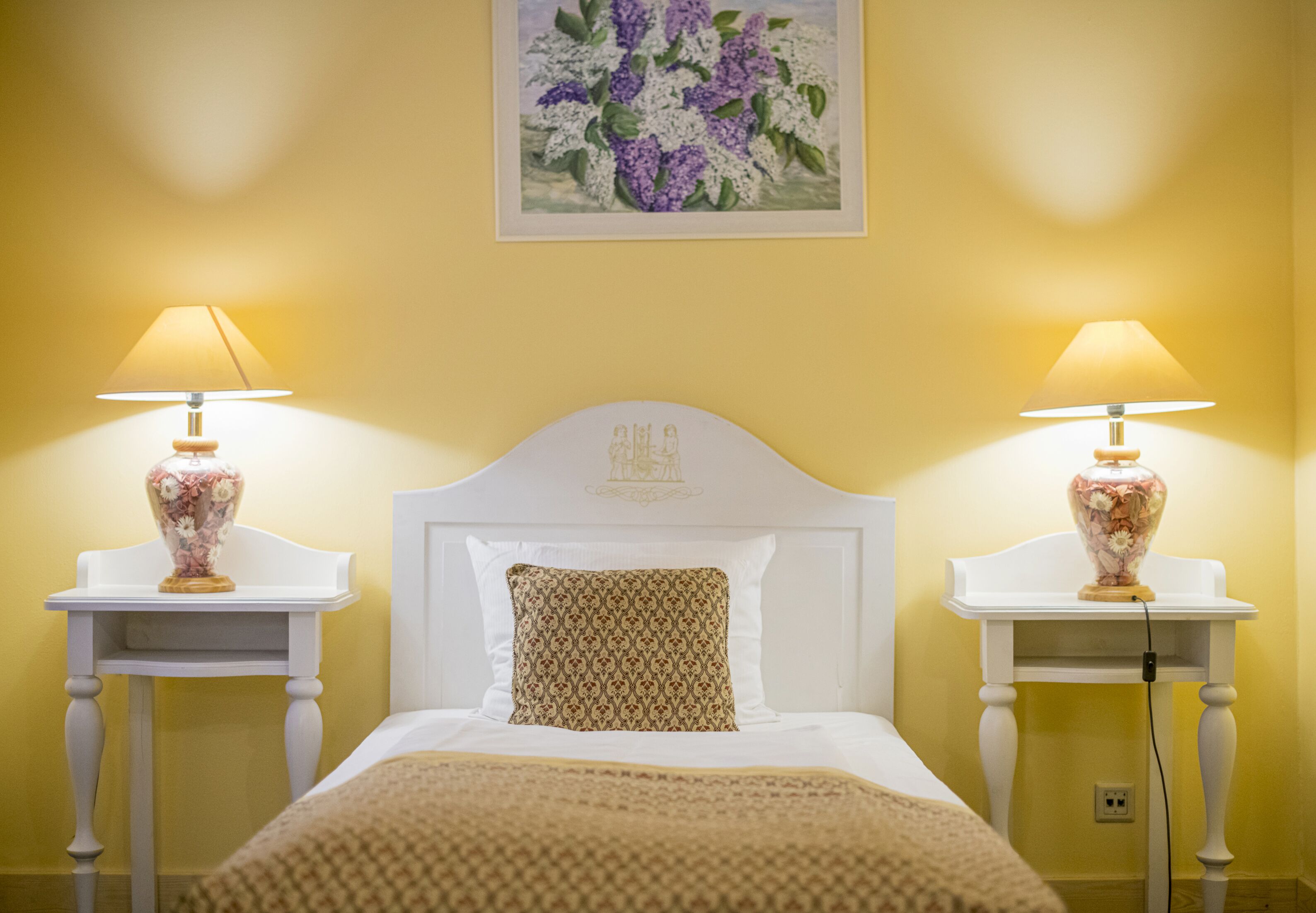 A neatly made bed with a patterned pillow, two bedside tables with lamps, and a painting of flowers on the wall.
