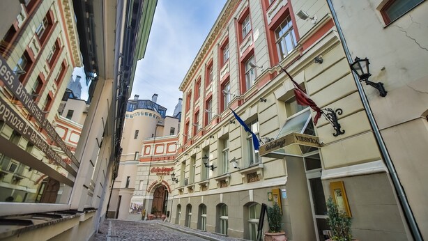 A cobblestone street with historic buildings, a prominent archway, and a sign reading "Viesnica." - Gutenbergs Hotel (Riga)