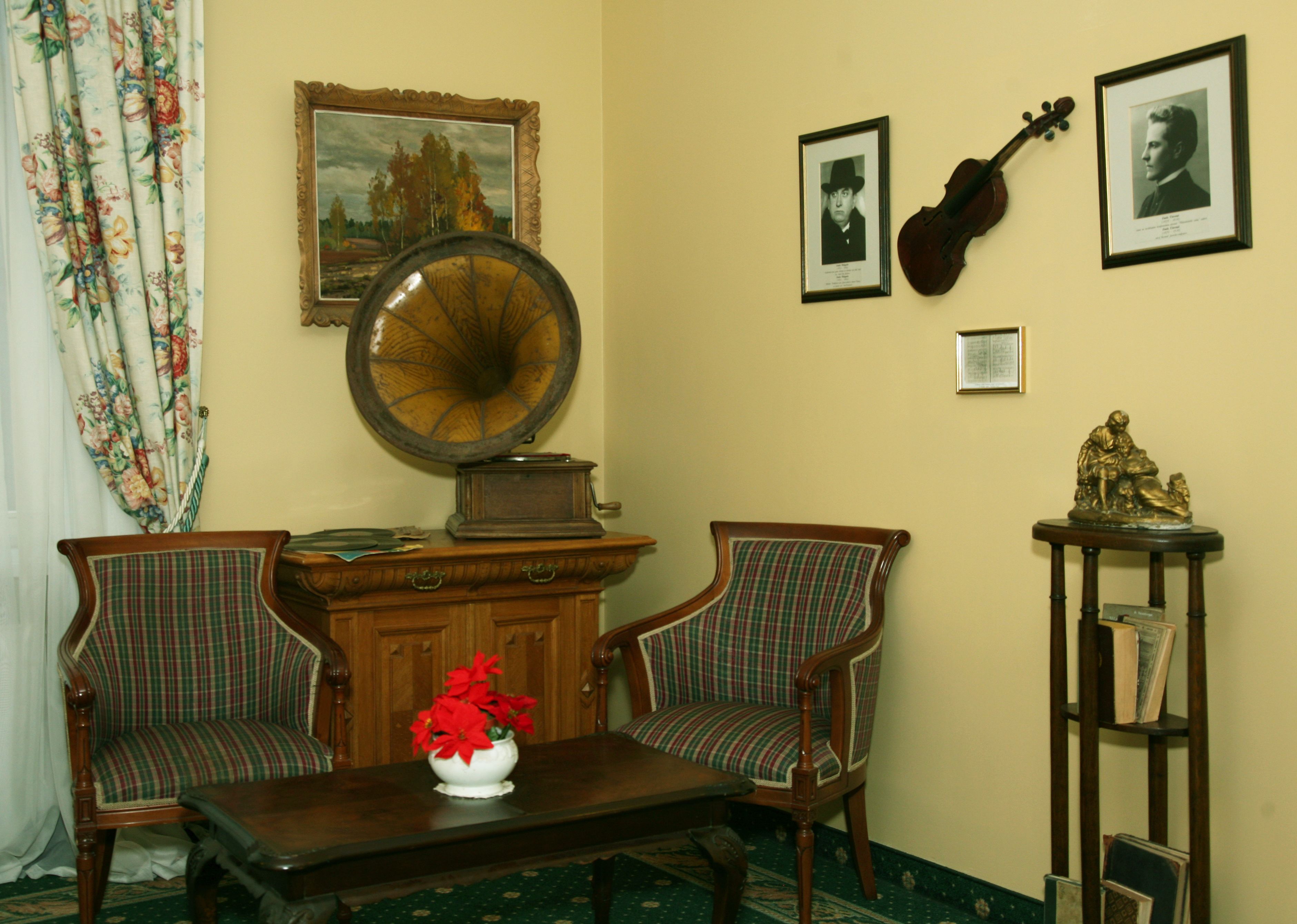 Una stanza con due poltrone imbottite, un tavolo in legno con un vaso di fiori rossi, un grammofono, quadri appesi, un violino e una statua su un tavolino.