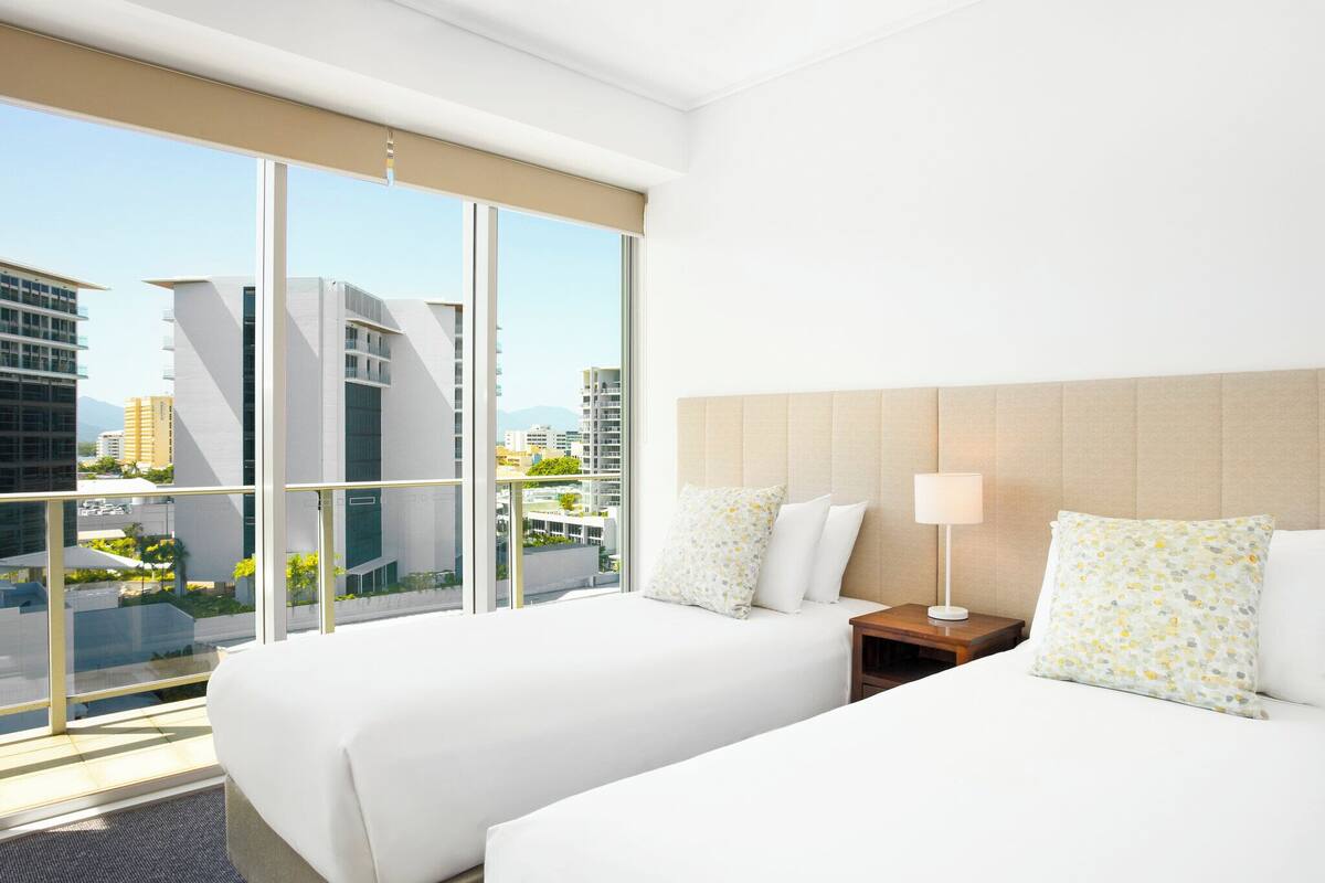 A hotel room with two beds, a nightstand, a lamp, and a large window offering a city view.