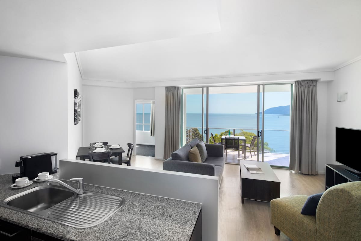 A modern hotel room with a kitchen area, a living room with a sofa and a TV, and a balcony with a view of the sea.