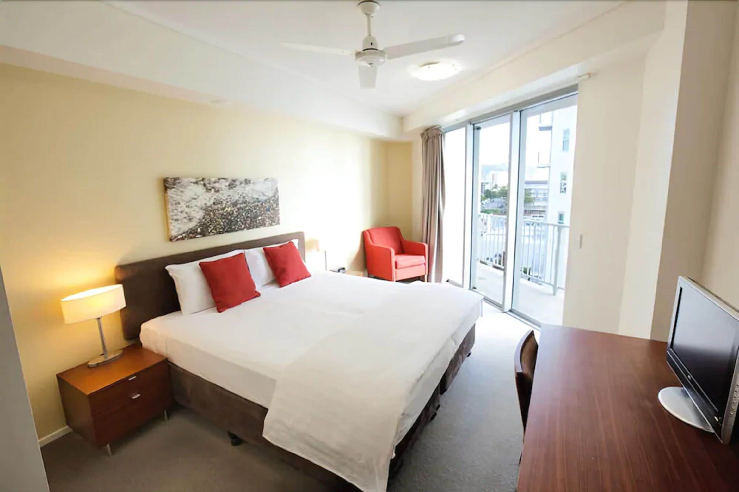 A hotel room with a bed, a TV, a desk, a chair, a ceiling fan, and a balcony with a view.