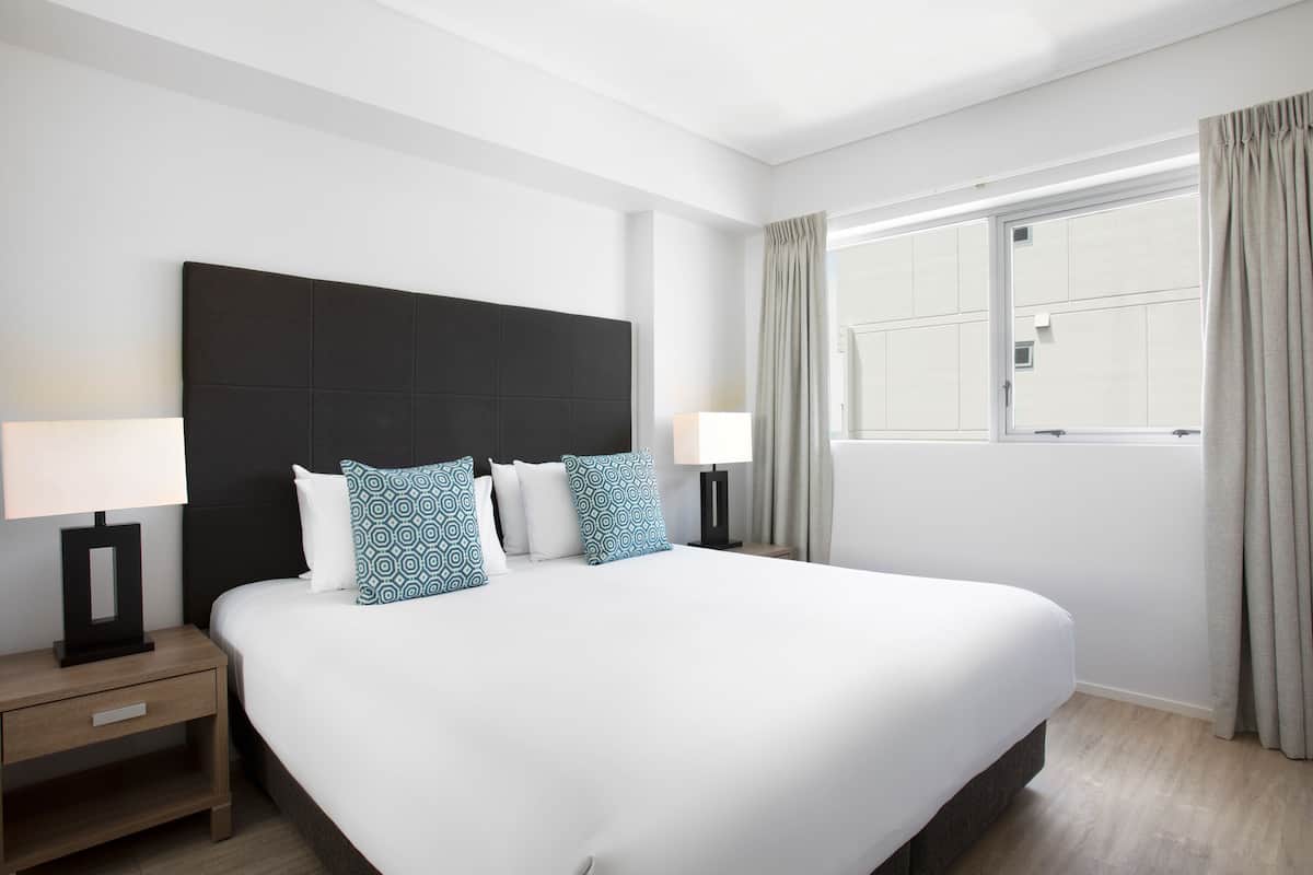 A large bed with a dark headboard, two bedside tables with lamps, patterned pillows, and a window with curtains.