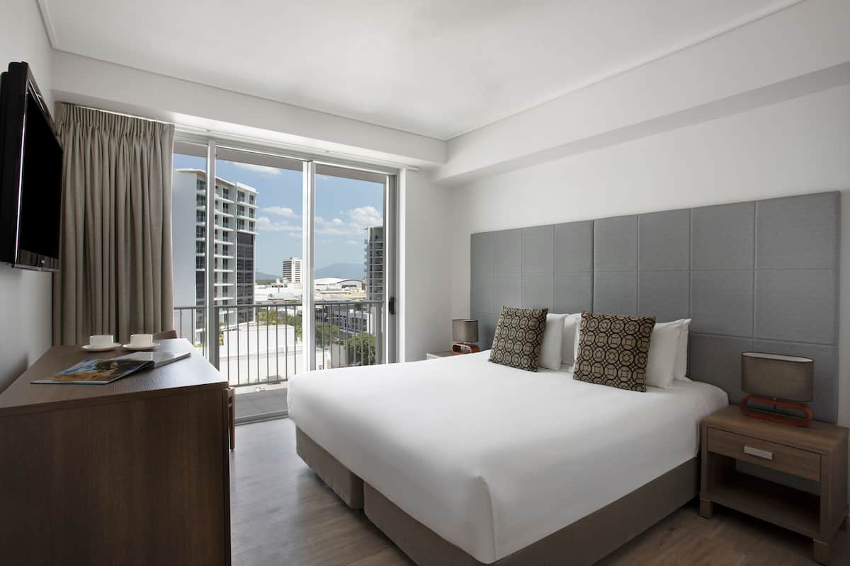 A modern hotel room with a large bed, a desk, a television, and a balcony with a city view.