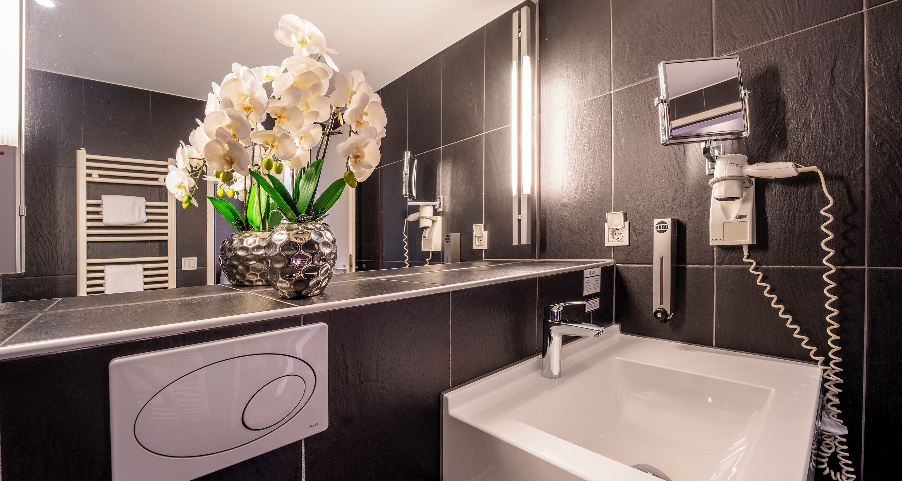 A modern bathroom with a white sink, a handheld showerhead, and a vase of white orchids.