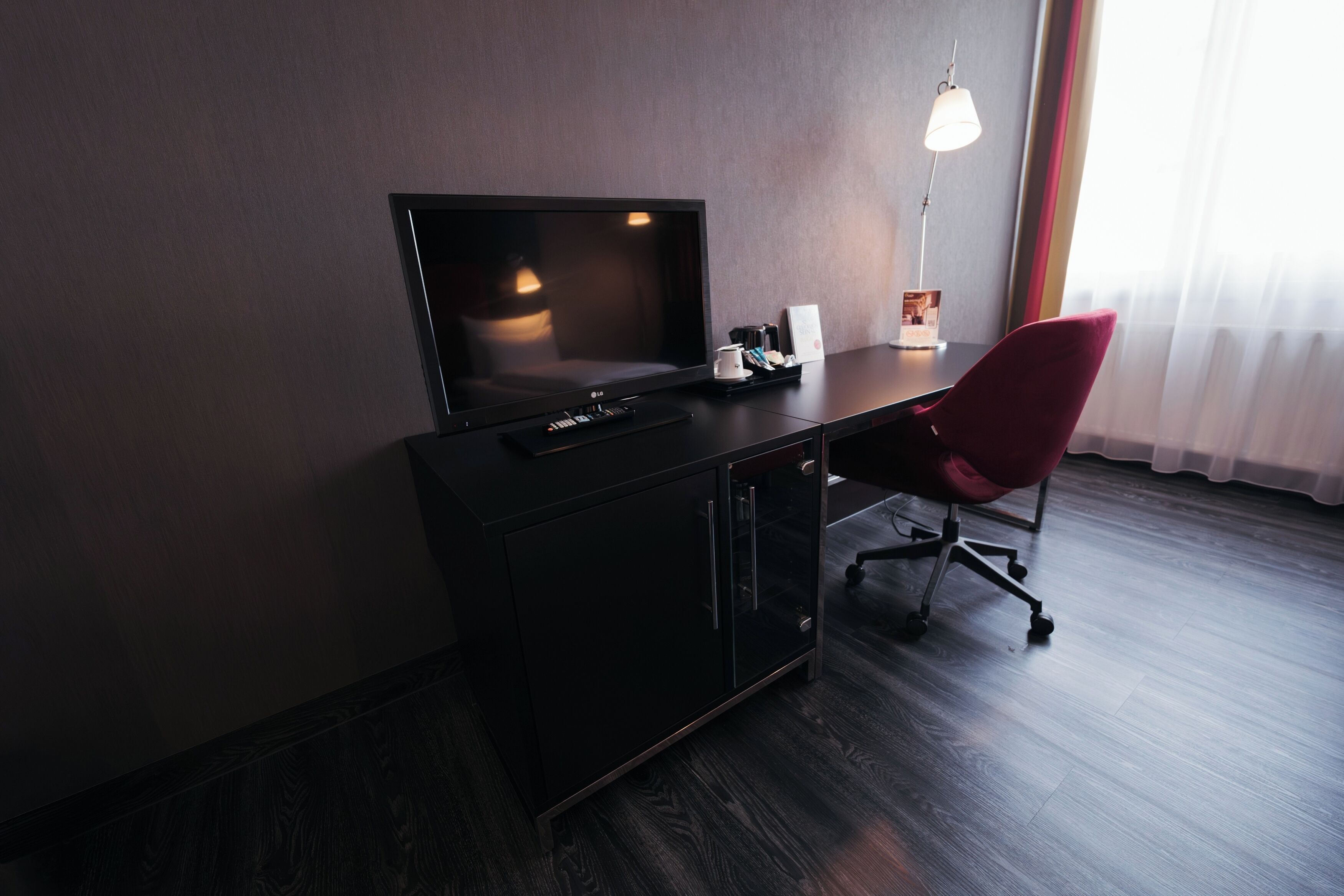 Una habitación de hotel con piso de madera oscura, un escritorio negro con televisor de pantalla plana, una silla roja, una lámpara de escritorio y una ventana con cortinas translúcidas.