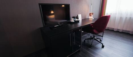 Una habitación de hotel con piso de madera oscura, un escritorio negro con televisor de pantalla plana, una silla roja, una lámpara de escritorio y una ventana con cortinas translúcidas.