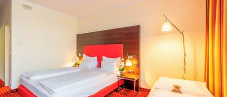 Habitación de hotel con cama, mesita de noche, lámpara y un oso de peluche sobre la cama.