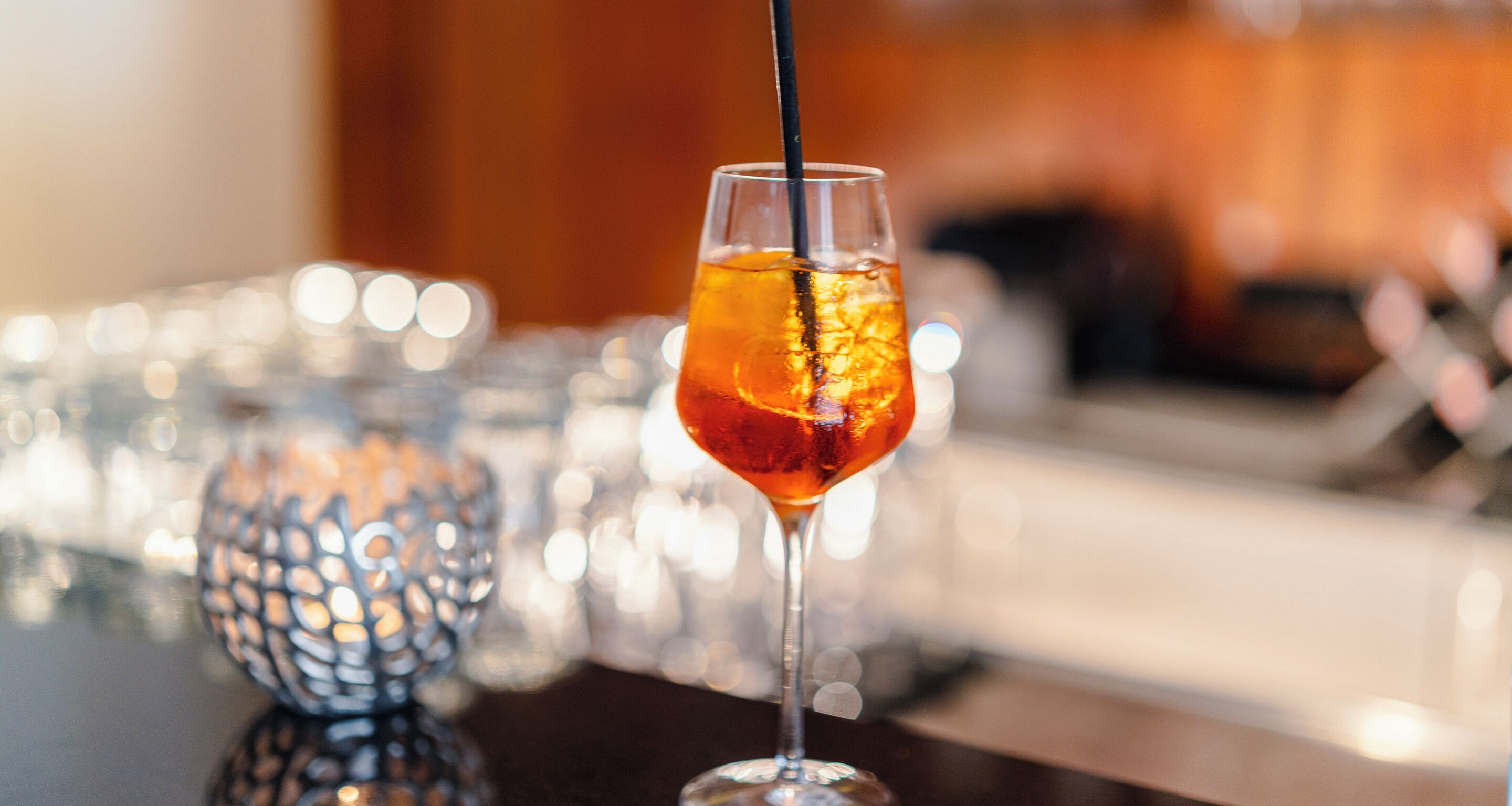 Un verre contenant une boisson de couleur ambrée et des glaçons, posé sur une surface réfléchissante, avec un arrière-plan flou mettant en scène un objet décoratif en métal.