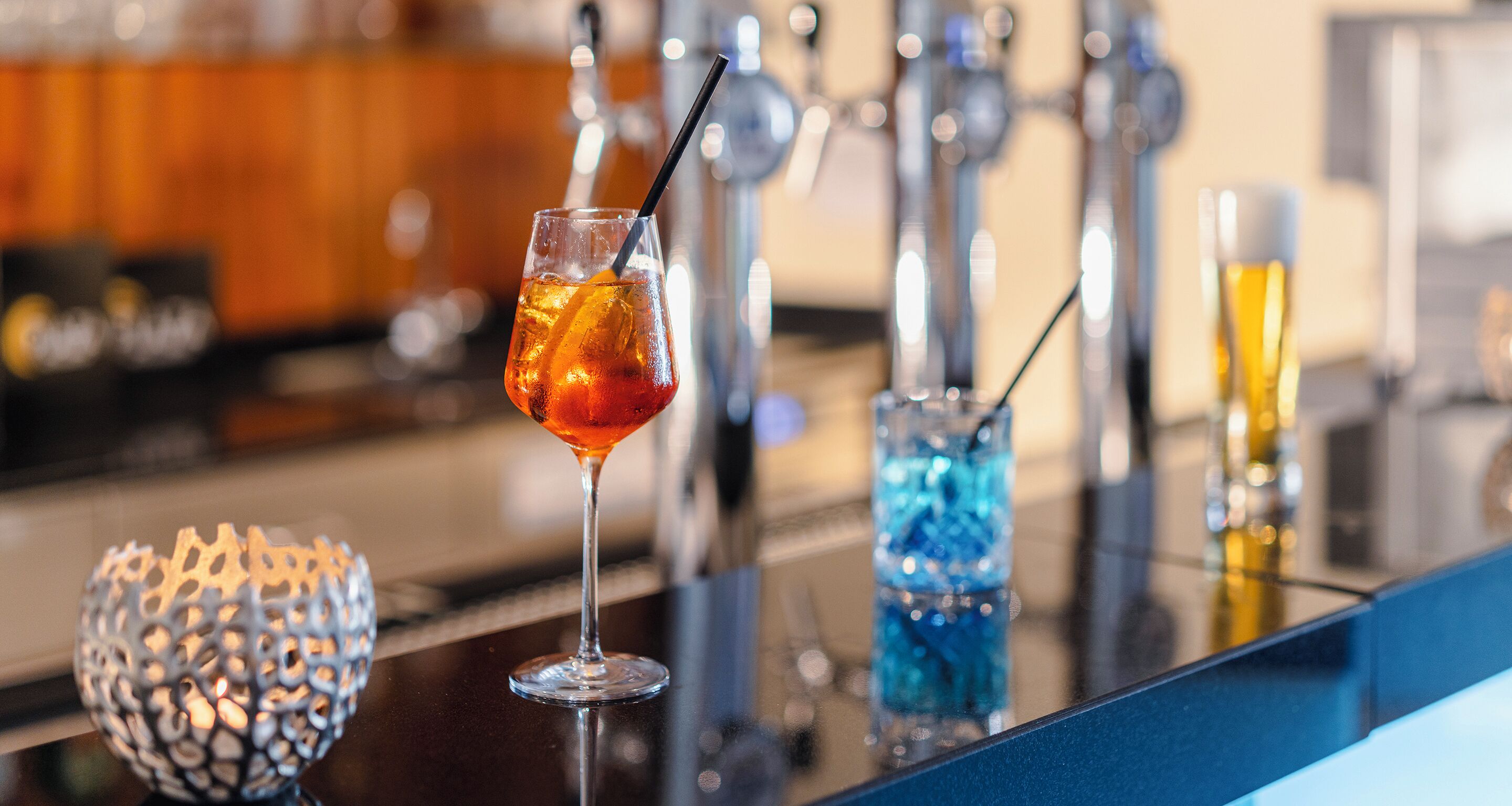 Een baromgeving met een glas amberkleurig drankje, een blauw decoratief glas en een kandelaar.